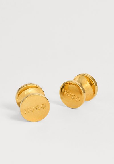 Paire de boutons de manchette ronds en or gravés avec le mot "HUGO" sur fond blanc.