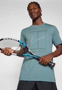 Τιρκουάζ κοντομάνικο μπλουζάκι με πολύχρωμο γραφικό λογότυπο Nike. Ο παίκτης κρατά μια μαύρη και μπλε τένις ρακέτα με υφή στην λαβή.