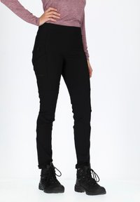 Black outdoor pants with a slim fit, featuring a high waist, zippered side pockets, and reinforced seams. Worn with black hiking boots.