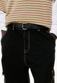 Pantaloni in velluto a coste neri con dettagli di cucitura bianca, abbinati a una maglia a righe beige e rosse. Una cintura nera con fibbia argentata completa il look.
