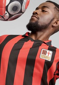 Zwarte en rode gestreepte voetbalshirt met een kraag, voorzien van een clubembleem op de linkerborst en een gestructureerde stof. Een zwart-witte voetbal is in beweging.