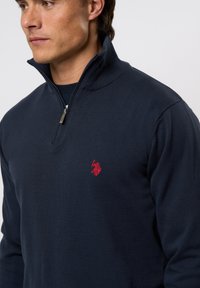 Fiatal férfi, sötét tengerészkék cipzáras pulóvert visel, bal mellkasán kis piros hímzett logóval, balra néz, egyszerű háttér előtt.