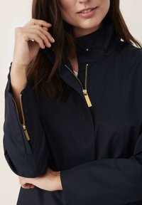 Marinejakke med højt krave, der har guld lynlåse foran og på ærmerne. Glat tekstur, afslappet design og minimalistische accenter.