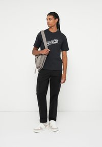 Tricou negru cu grafică albă, asociat cu pantaloni cargo negri și adidași bej. Modelul poartă o geantă gri de umăr.