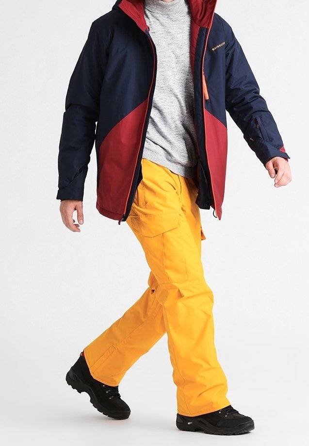 Personne marchant portant une veste bleu marine et rouge, une chemise grise, un pantalon jaune vif et des chaussures de randonnée noires.