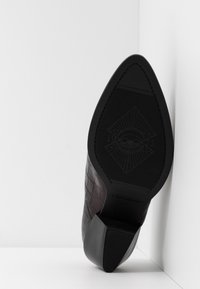 Mörkbrun, strukturerad högklackad sko sedd från botten, som visar upp sin spetsiga tå och märkta sula mot en vit bakgrund.