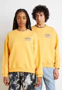 Sárga pulóverek, amelyek elülső mellkasi logóval, bordázott mandzsettákkal és szegélyekkel rendelkeznek. Az egyik modellen tye-dye nadrág van; a másikon világoskék nadrág.