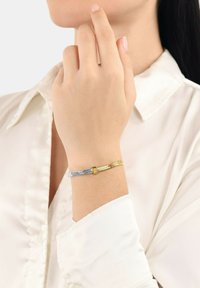Bracciale in metallo con toni intrecciati d'argento e d'oro, con inserti rettangolari e superficie testurizzata, indossato su un polso nudo contro una camicia chiara.