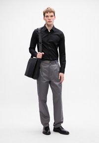 Zwarte overhemd met knopen, grijze broek en zwarte leren schoenen. Een zwarte schoudertas met een schouderriem wordt in de rechterhand vastgehouden.