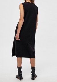 Abito nero senza maniche con una vestibilità rilassata, lunghezza al ginocchio, colletto a righe, spacco laterale e texture liscia; abbinato a stivali neri alla caviglia.