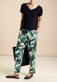 Kvinna klädd i en svart V-ringad kortärmad topp, vita byxor med stort grönt lövtryck, svarta mesh-lägenheter och håller en svart väska.