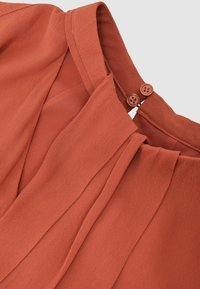 Terrakota bluzica z gladko teksturo, z nagubaním in zapenjanjem na hrbtu z dvema rjavima gumboma.