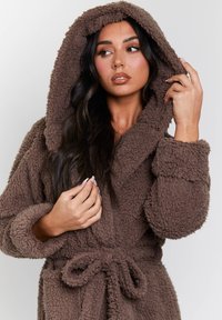 Accappatoio marrone, morbido e pelooso, con cappuccio, maniche lunghe e vita regolabile. Texture soffice con una leggera lucentezza. Presenta ampi polsini accoglienti.