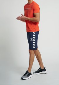 Hombre con camiseta de manga corta naranja y pantalones cortos deportivos azul marino con rayas en forma de chevron blancas, de pie y aplaudiendo.