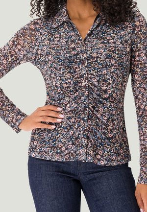 Femme portant un chemisier ajusté à manches longues à fleurs roses, bleues et noires, associé à un jean bleu foncé, main sur la hanche.