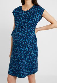 Sukienka ciążowa w ciemnoniebieskim kolorze z geometrycznym wzorem w jasnoniebieskim. Krótkie rękawy, luźny krój z regulowanym pasem. Gładka tekstura materiału.