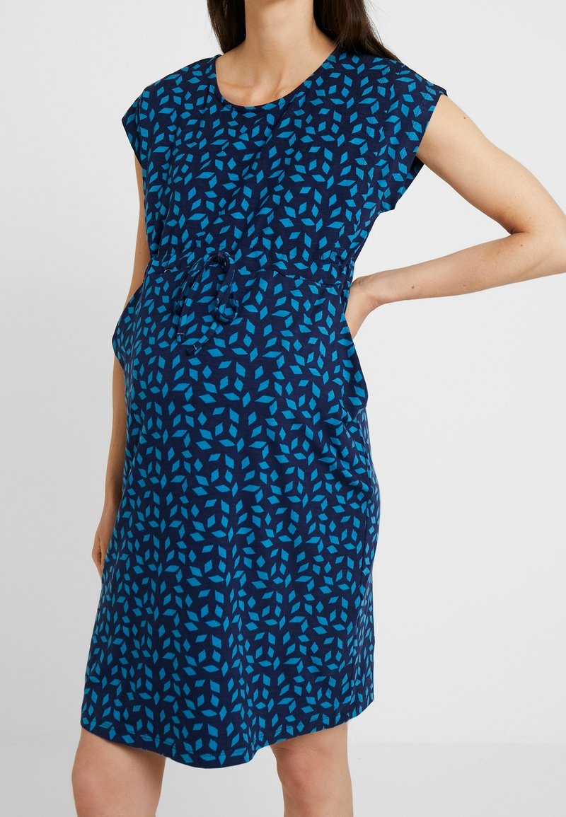 Sukienka ciążowa w ciemnoniebieskim kolorze z geometrycznym wzorem w jasnoniebieskim. Krótkie rękawy, luźny krój z regulowanym pasem. Gładka tekstura materiału.