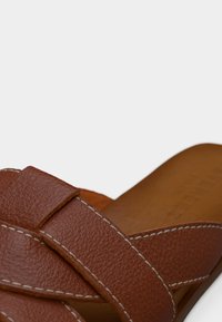 Καφέ δερμάτινα σανδάλια με σχέδιο σταυρωτών λουριών, υφή στην επιφάνεια, αντίθετη λευκή ραφή και επίπεδη σόλα.