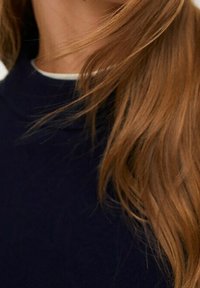 Suéter navy con un cuello alto, que presenta un borde blanco. El cabello castaño es parcialmente visible, mostrando una textura suave y ondulada.