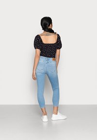 Blouse noire à motifs floraux avec des manches bouffantes, assortie à un jean skinny bleu clair ; stylisée avec des baskets blanches. L'arrière-plan est uni.