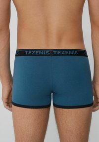 Boxer de hombre en color verde azulado sólido, fabricados en tela suave, con una cinturilla elástica negra que presenta la marca TEZENIS. Diseño de pierna corta.