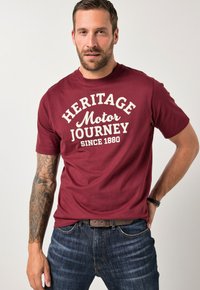 Maroonfarbenes Baumwoll-T-Shirt mit dem cremefarbenen Schriftzug "HERITAGE Motor JOURNEY SINCE 1880", Rundhalsausschnitt, kurze Ärmel, kombiniert mit dunkelblauen Jeans.