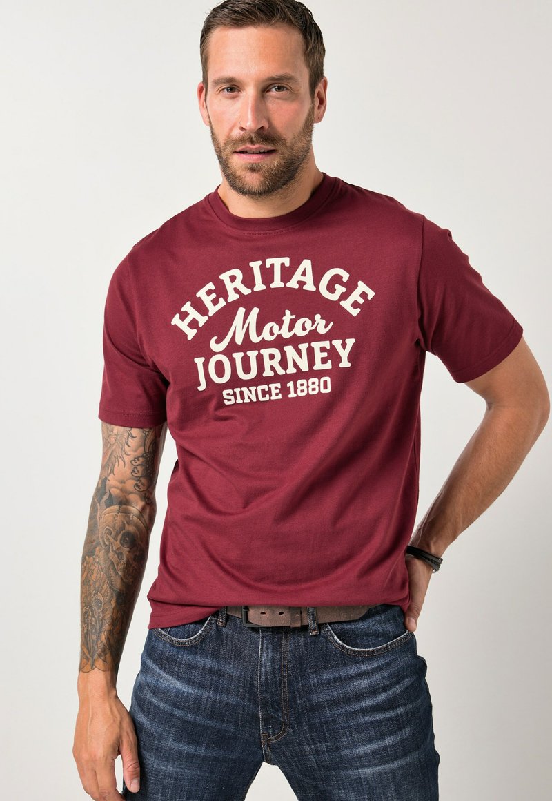 Maroonfarbenes Baumwoll-T-Shirt mit dem cremefarbenen Schriftzug "HERITAGE Motor JOURNEY SINCE 1880", Rundhalsausschnitt, kurze �Ärmel, kombiniert mit dunkelblauen Jeans.
