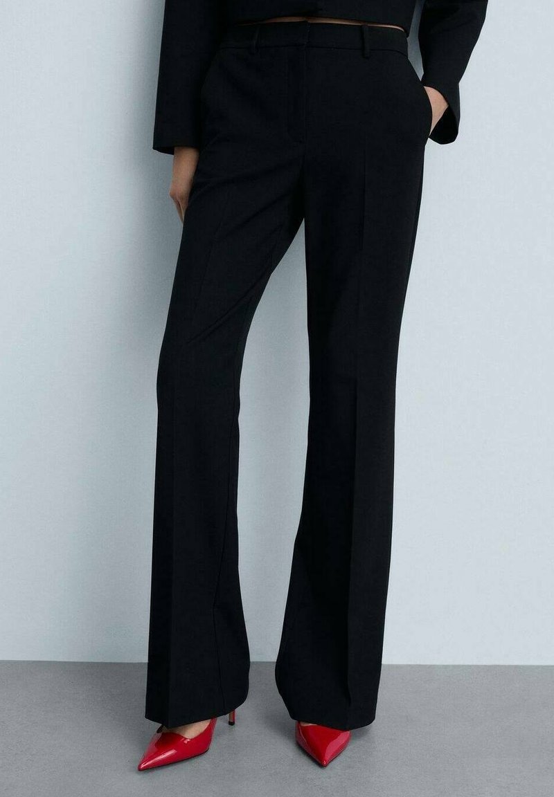 Personne portant un pantalon noir taille haute à jambes larges avec une ceinture marron et des escarpins rouges à bouts pointus, debout devant un fond uni.