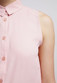 Ärmløs lyserød bluse med krave og knaplukning fortil, fremstillet af glat stof og små, runde knapper.