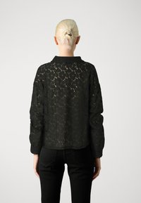 Blusa de renda preta com padrão floral, apresentando mangas longas e colarinho. O design tem uma textura semi-transparente. Usada sobre calças pretas.