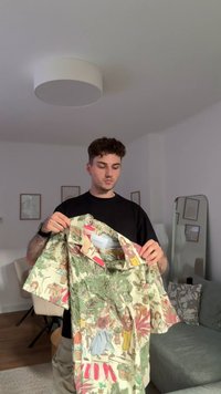 Korte mouwen shirt met een kleurrijk, geïllustreerd patroon van mensen te midden van groen op een lichte achtergrond. De stof lijkt lichtgewicht en glad te zijn.