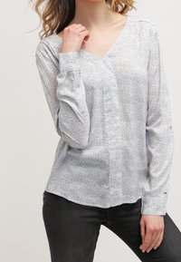 Biała, długa bluzka z długim rękawem i dekoltem w serek, ozdobiona rozproszonym wzorem czarnych kropek. Gładka faktura, guziki na rękawach, dopasowany krój.