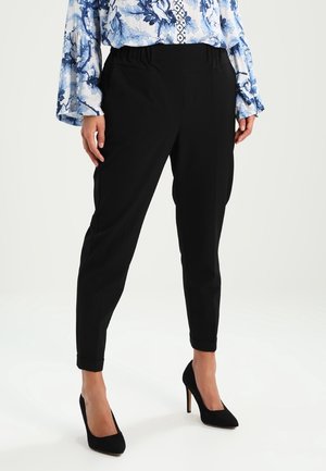 Pantalon noir fuselé avec une texture lisse, taille élastique et coupe taille cheville, associé à des chaussures à talons hauts noires.