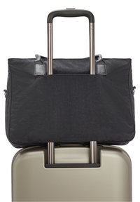 Sac de voyage noir posé sur la poignée déployée d'une valise rigide beige à roulettes sur un fond blanc.