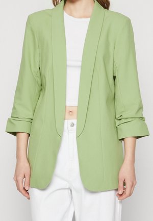 Persona que lleva un blazer abierto de color verde claro con las mangas remangadas, sobre un top corto blanco y pantalones blancos de talle alto.