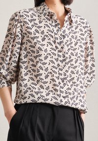 Skjorta i ljus cream med svart bladmönster. Har en klassisk krage och lösa ärmar, matchas med skräddarsydda svarta byxor.