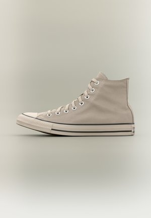 Zapatilla alta de lona color beige con suela y puntera de goma blanca, cordones blancos y ojales metálicos, mostrada de perfil sobre un fondo neutro.