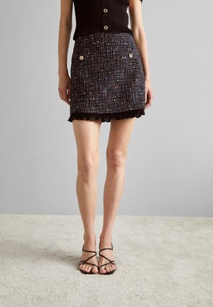 Zwart tweed minirok met kleurrijke vlekken, heeft gouden knoopdetails en een gerimpelde zoom. Gecombineerd met zwarte bandjesandalen.