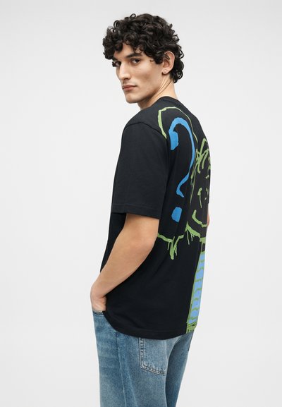 Jeune homme aux cheveux bouclés portant un t-shirt noir à motif graphique avec un design abstrait bleu et vert, les mains dans les poches, de profil.