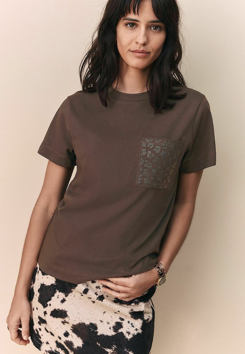 T-shirt marron à manches courtes avec une poche en imprimé animal texturé. Le modèle le porte avec une jupe à motifs noir et blanc, présentant un look décontracté.