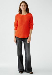 Roter gerippterPullover mit weitem Halsauschnitt und langen Ärmeln, kombiniert mit dunklen Schlagjeans. Das Outfit zeichnet sich durch einen lockeren Sitz und eine subtile Textur aus.