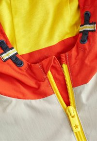 Farverig jakke med en gul hætte, orange krave og cremefarvet krop med en gul lynlås og sorte snore. Tekstureret stof.