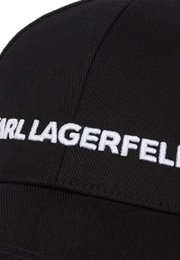 Fekete sapka texturált anyagból, fehér hímzett "KARL LAGERFELD" felirattal az elején. Görbült karimával és struktúrált dizájnnal.