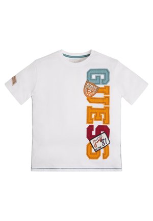 T-shirt blanc à manches courtes avec des lettres verticales colorées "GUESS" et des patches sur le côté droit avant et un patch sur la manche gauche.