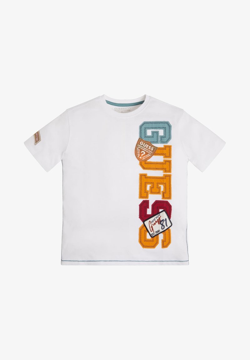 T-shirt blanc à manches courtes avec des lettres verticales colorées "GUESS" et des patches sur le côté droit avant et un patch sur la manche gauche.