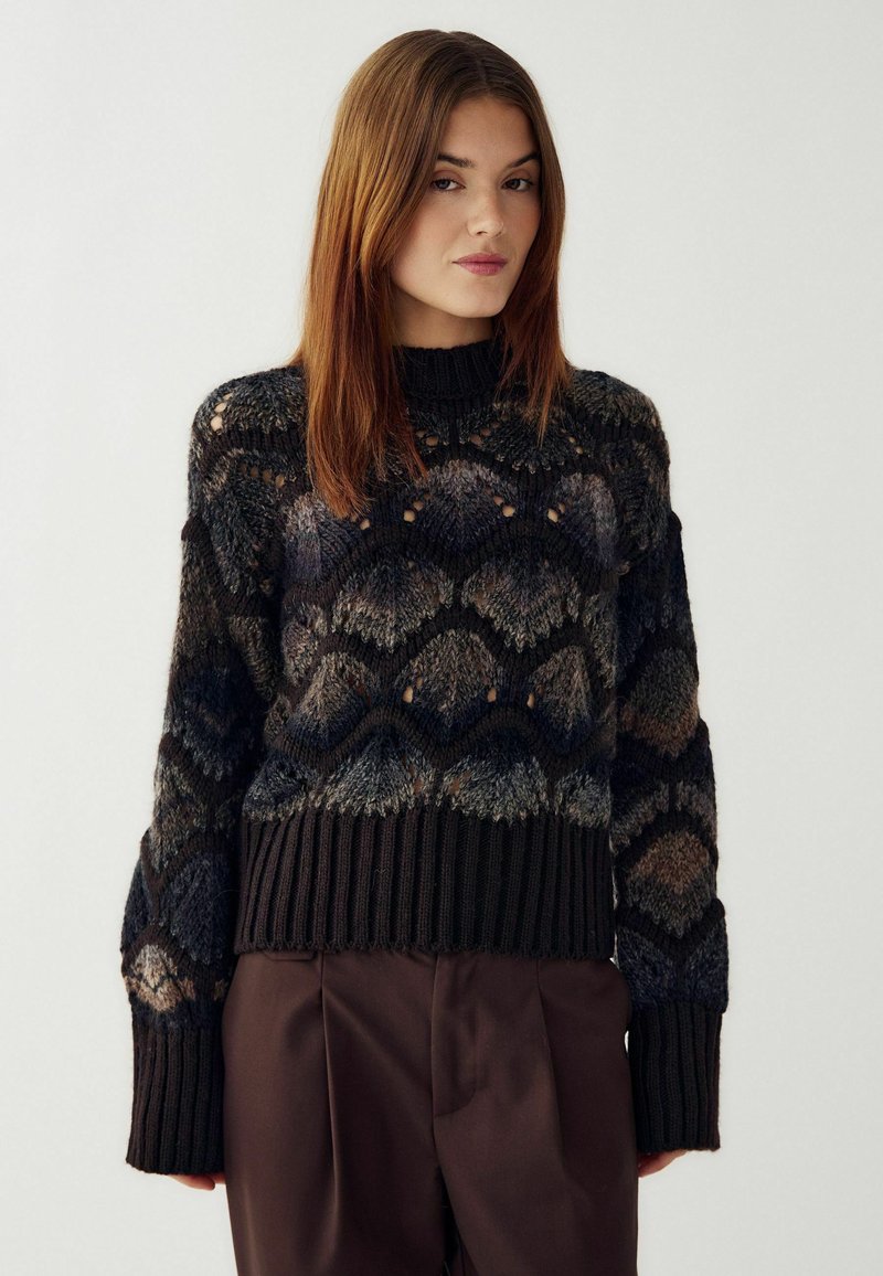 Megzta megztinis su geometriniu raštu tamsiomis spalvomis. Turi aukštą kaklą, plačias rankoves ir smulkiai raštuotą apatinę dalį, pagamintas iš tekstūruotos siūlų.