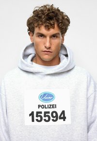 Grijze hoodie met een groot wit label waarop "POLIZEI 15594" staat. Krullend haar, neutrale achtergrond, met focus op de textuur en ontwerpelementen van de hoodie.