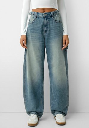 Persoon draagt loszittende vervaagde blauwe spijkerbroek, wit crop top met lange mouwen en witte sneakers met rubberen zolen, staand tegen een eenvoudige muur.