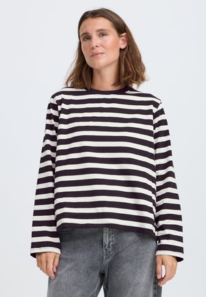 Femme aux cheveux bruns mi-longs, vêtue d'un t-shirt à manches longues rayé noir et blanc et d'un jean gris, debout devant un fond uni.