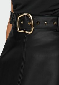 Gros plan sur une jupe en cuir noir avec une large ceinture noire dotée d'une grande boucle carrée en or et d'œillets dorés, portée avec un haut noir.
